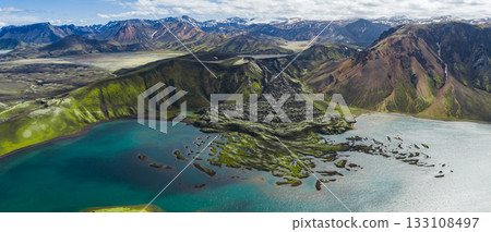 Aerial view of a turquoise lake in Iceland's Landmannalaugar region, surrounded by mossy lava fields, multicolored mountains, and snow capped peaks. 133108497