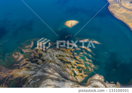 Aerial image of a deep blue lake in Iceland featuring a small island, visible underwater patterns, curved shoreline, and rocky terrain with moss. 133108501