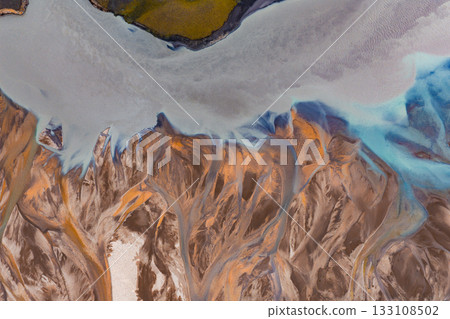 Aerial image of a braided river system in Iceland, featuring orange, brown, and blue hues with green vegetation at the top of the landscape. 133108502
