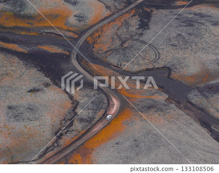 An aerial view shows a dirt road winding through Iceland's volcanic terrain, with dark ashen patches, orange and yellow hues, and a single white vehicle. An aerial view shows a dirt road winding through Iceland's volcanic terrain, with dark ashen patches, orange and yellow hues, and a single white vehicle. 133108506