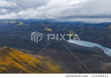 A dramatic aerial view of Iceland's volcanic terrain with dark rocks, yellow moss, a pale blue lake, winding rivers, scattered hills, and overcast skies. A dramatic aerial view of Iceland's volcanic terrain with dark rocks, yellow moss, a pale blue lake, winding rivers, scattered hills, and overcast skies. 133108512