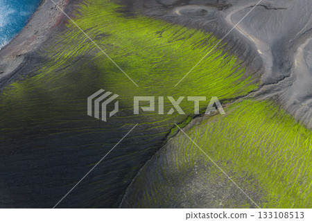 Aerial view of vibrant green moss on dark volcanic soil in Iceland, with rippling textures and a blue water body in the upper left corner. Aerial view of vibrant green moss on dark volcanic soil in Iceland, with rippling textures and a blue water body in the upper left corner. 133108513