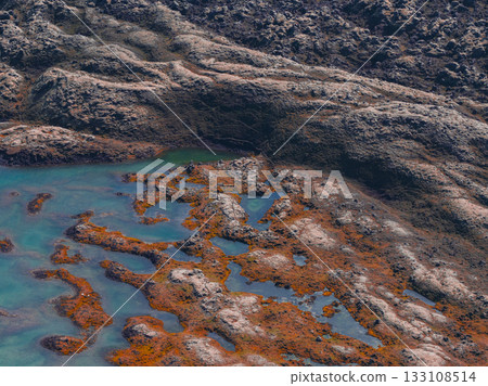 Rocky terrain with shallow turquoise pools, orange and brown moss, and dark textured rocks, showcasing Iceland's volcanic and geothermal features. 133108514