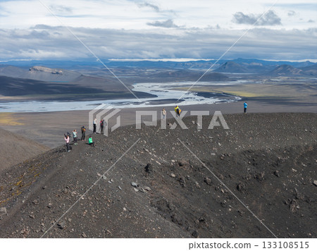 A group of people walks on a volcanic crater rim in Iceland, surrounded by rocky terrain, a winding river, distant mountains, and a cloudy sky. 133108515