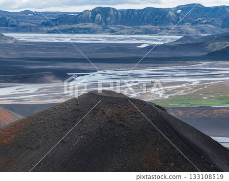 A dark volcanic hill with reddish hues stands before braided rivers on a vast plain, framed by rugged snow streaked mountains in Iceland. 133108519