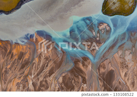 Braided blue glacial rivers in Iceland flow through earthy brown and orange terrain, with green moss covered land visible in the upper right corner. Braided blue glacial rivers in Iceland flow through earthy brown and orange terrain, with green moss covered land visible in the upper right corner. 133108522