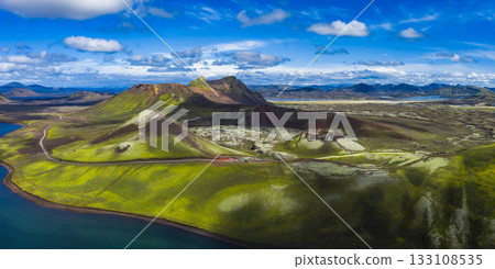Aerial view of Iceland's moss covered hills, volcanic craters, and a serene lake under a partly cloudy sky, showcasing diverse textures and colors. 133108535