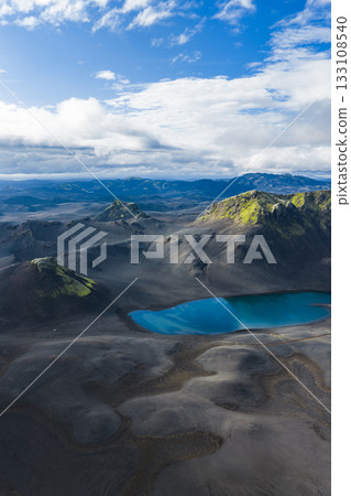 A vivid blue crater lake sits amid dark volcanic terrain in Iceland, with moss covered hills and mountains under soft sunlight and a partly cloudy sky. 133108540