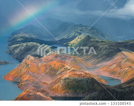 Aerial view of Iceland's rainbow mountains with multicolored terrain, a vivid rainbow in the sky, and serene lakes nestled among the hills. Aerial view of Iceland's rainbow mountains with multicolored terrain, a vivid rainbow in the sky, and serene lakes nestled among the hills. 133108543