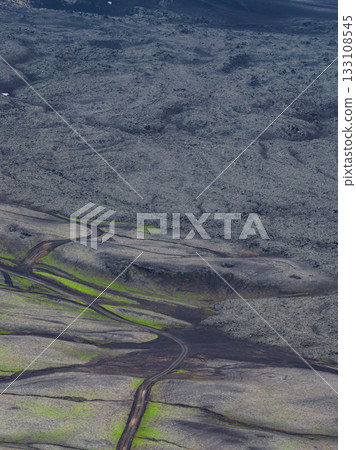 An aerial view of Iceland's rugged volcanic landscape, featuring dark terrain, vibrant green moss patches, and a winding dirt road cutting through. 133108545
