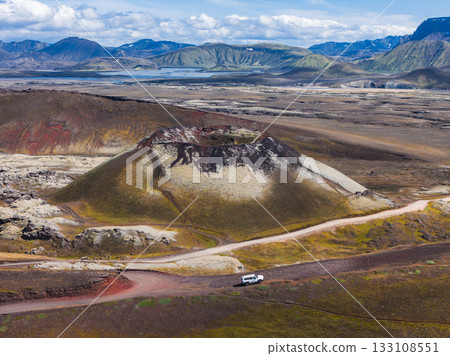 A volcanic crater in Iceland surrounded by red, brown, and green terrain. A dirt road with a white vehicle encircles the crater, with hills and a lake nearby. 133108551