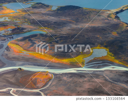 Aerial view of glacial rivers winding through dark volcanic terrain in Iceland, with vibrant vegetation and scattered lakes reflecting the sky. 133108564