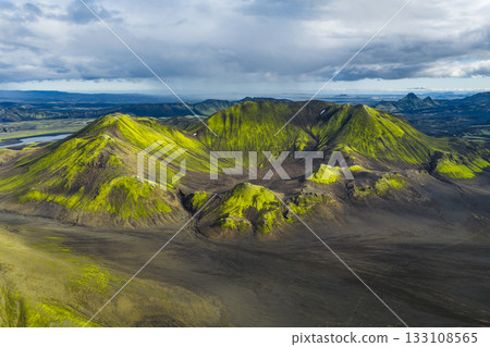 Aerial view of green moss covered volcanic hills surrounded by dark terrain in Iceland, with a winding river and distant mountains under a cloudy sky. Aerial view of green moss covered volcanic hills surrounded by dark terrain in Iceland, with a winding river and distant mountains under a cloudy sky. 133108565