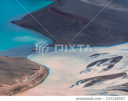 A vivid turquoise lake surrounded by dark volcanic terrain with visible sediment patterns, light deposits, and intricate ridges in Iceland. A vivid turquoise lake surrounded by dark volcanic terrain with visible sediment patterns, light deposits, and intricate ridges in Iceland. 133108567