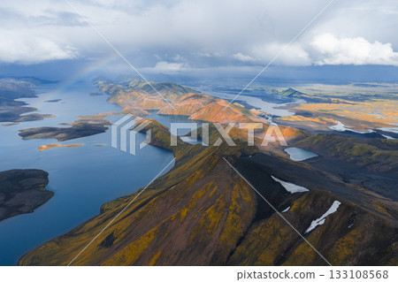 Aerial view of Icelandic highlands with vibrant rhyolite mountains, winding lakes, patches of snow, mossy greens, and a faint rainbow in the sky. 133108568