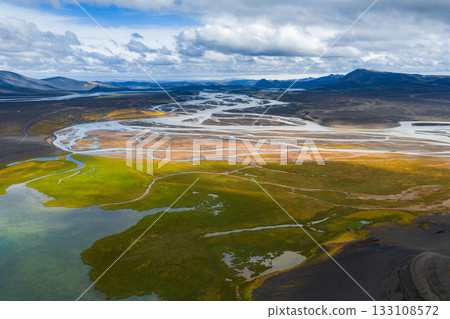 Aerial view of braided rivers flowing through a plain with green and golden moss, volcanic soil, and distant mountains under a partly cloudy sky. 133108572