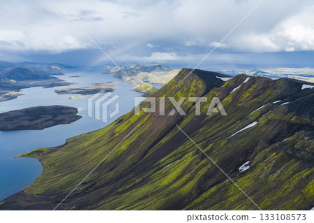 Aerial view of green and black volcanic mountains with snow patches, a serene lake with islands, and a faint rainbow under a dramatic sky in Iceland. Aerial view of green and black volcanic mountains with snow patches, a serene lake with islands, and a faint rainbow under a dramatic sky in Iceland. 133108573