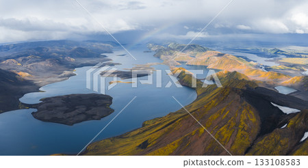 A vast lake in Iceland surrounded by rugged volcanic terrain with yellow and orange hills, deep blue water, and a faint rainbow under a cloudy sky. 133108583
