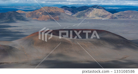 A volcanic crater with reddish and gray tones surrounded by barren ash covered land, multicolored mountains, and a winding road under a cloudy sky. A volcanic crater with reddish and gray tones surrounded by barren ash covered land, multicolored mountains, and a winding road under a cloudy sky. 133108586