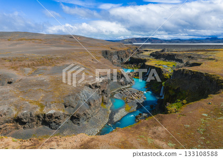 Sigoldugljufur Canyon in Iceland features turquoise waters, dark volcanic rock, small waterfalls, mossy patches, and a partly cloudy sky. 133108588