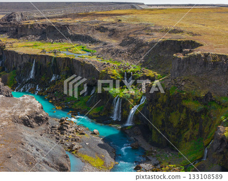 Turquoise river winds through Sigoldugljufur Canyon in Iceland, surrounded by moss covered cliffs, cascading waterfalls, and a barren plateau above. 133108589