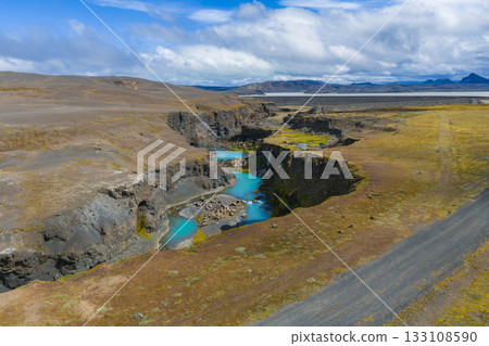 Sigoldugljufur Canyon in Iceland features turquoise streams, dark volcanic rocks, green moss, a dirt road, and distant mountains under a cloudy sky. 133108590