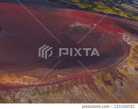 Aerial view of a volcanic crater in Iceland, featuring vivid red and orange hues, green moss, dark soil, and rugged terrain with vibrant vegetation. 133108592