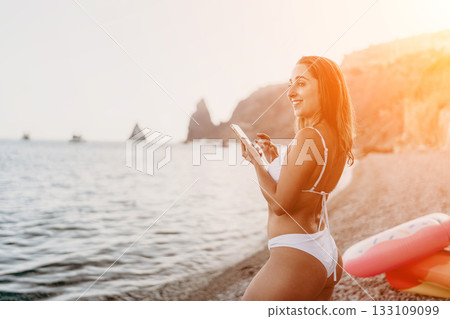 Woman, smartphone, beach. Smiling woman in bikini texts on phone at pebble beach with copy space. Woman, smartphone, beach. Smiling woman in bikini texts on phone at pebble beach with copy space. 133109099
