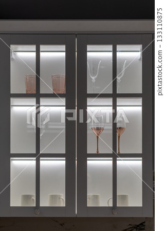 A close-up shot of a white kitchen cabinet with glass doors and internal LED lighting A close-up shot of a white kitchen cabinet with glass doors and internal LED lighting 133110875