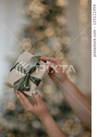 Hands Holding Gift Box with Satin Bow by Christmas Tree 133111459