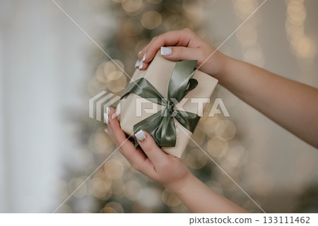 Hands Holding Gift Box with Satin Bow by Christmas Tree Hands Holding Gift Box with Satin Bow by Christmas Tree 133111462
