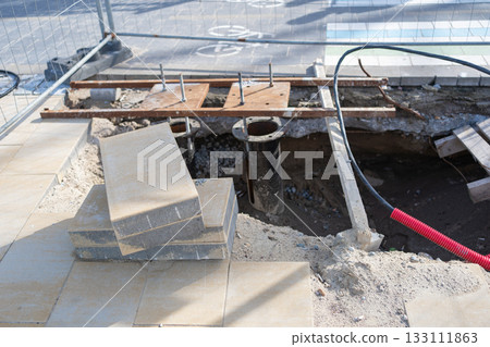 Open construction pit with pipes, boards and removed paving slabs. Documenting infrastructure work, underground utility installation and urban engineering processes in detail 133111863