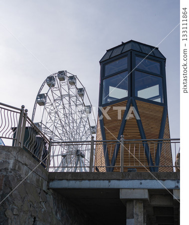 Ferris wheel next to a tall wooden observation tower against a partly cloudy sky. A combination of modern entertainment and architectural structures in an urban setting Ferris wheel next to a tall wooden observation tower against a partly cloudy sky. A combination of modern entertainment and architectural structures in an urban setting 133111864
