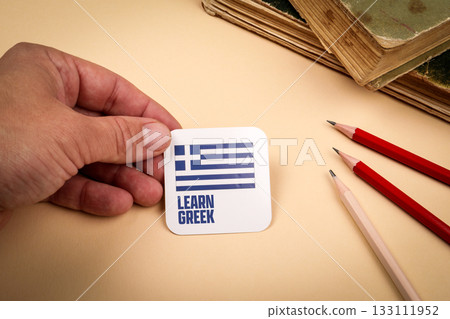 Hand holding card with Greek flag and text learn Greek on beige background Hand holding card with Greek flag and text learn Greek on beige background 133111952