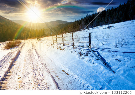 winter solstice in mountainous countryside. day and night time change concept. wooden fence along the road through coniferous forest near snow covered hills. scenic view of scene with sun and moon winter solstice in mountainous countryside. day and night time change concept. wooden fence along the road through coniferous forest near snow covered hills. scenic view of scene with sun and moon 133112205
