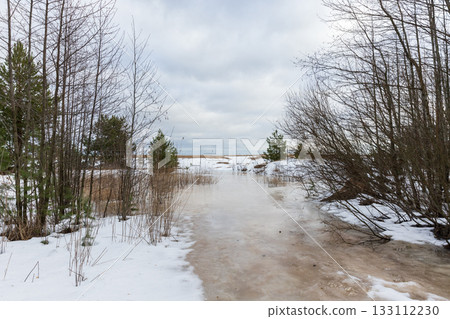 A quiet winter landscape shows an icy path winding through a snow-dusted forest 133112230