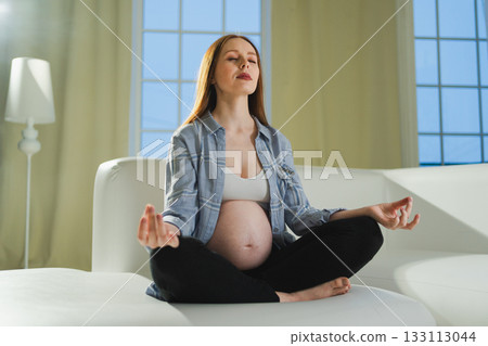 Pregnancy prenatal breathing. Pregnant woman with big belly sitting in lotus pose on sofa at home. Pregnant girl practicing yoga mindfulness meditation. Relaxation self care during pregnancy Pregnancy prenatal breathing. Pregnant woman with big belly sitting in lotus pose on sofa at home. Pregnant girl practicing yoga mindfulness meditation. Relaxation self care during pregnancy 133113044