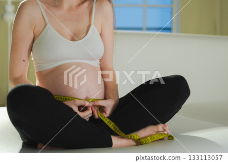 Pregnancy motherhood. Pregnant woman measuring big belly. Girl measuring her belly with tape to keep track of her fetus development. Woman enjoying pregnancy. Maternity tenderness parenthood new life Pregnancy motherhood. Pregnant woman measuring big belly. Girl measuring her belly with tape to keep track of her fetus development. Woman enjoying pregnancy. Maternity tenderness parenthood new life 133113057