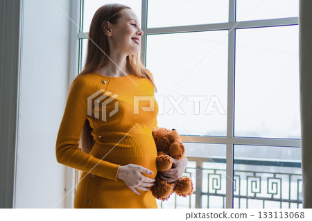 Pregnancy motherhood. Pregnant woman with big belly hugging teddy bear standing near window at home. Happy smiling girl enjoying pregnancy. Maternity tenderness parenthood new life concept 133113068
