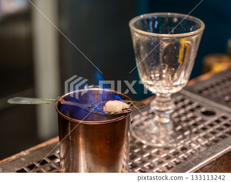 Prague, Czech Republic, August 8, 2023. A choreographic preparation of absinthe. A sugar cube soaked in the alcoholic beverage is burned on a special spoon, engulfed in a blue flame. Prague, Czech Republic, August 8, 2023. A choreographic preparation of absinthe. A sugar cube soaked in the alcoholic beverage is burned on a special spoon, engulfed in a blue flame. 133113242