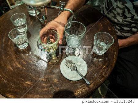 Prague, Czech Republic, August 8, 2023. A table with an absinthe still and glasses of water. A hand holds the glass of the green liqueur, alongside a special spoon and sugar cube. 133113243