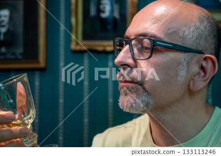 Prague, Czech Republic, August 8, 2023. Portrait of a middle-aged man drinking absinthe, looking into his glass. Relaxation and meditation in a bohemian setting. Artistic lifestyle. 133113246