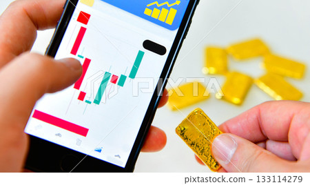 Hand holding a smartphone and a gold bar to check the gold price graph for investment Hand holding a smartphone and a gold bar to check the gold price graph for investment 133114279
