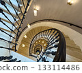 Spiral staircase viewed from above with ornate iron railing and warm hanging lights in historic interior 133114486