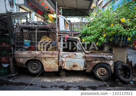 Old rusty wrecked car with trunk full of stuff parked outside building at alley corner in Bangkok. 133117021