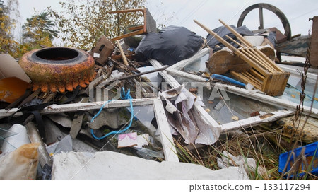 collection of industrial scraps and household junk at urban edge; corrugated panels, glass frames, metal containers and large wheel create textured composition against overgrown verge 133117294