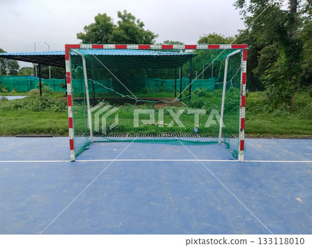 Outdoor futsal goalpost with striped metal frame and loose green net on blue sports court, standalone mini football goal in open ground surrounded by grass and trees, dedicated small-sided play area 133118010