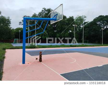 Empty outdoor basketball court with pink and blue painted surface, lone hoop post in quiet sports complex surrounded by greenery and tall light poles, peaceful athletic ground for games and recreation 133118018
