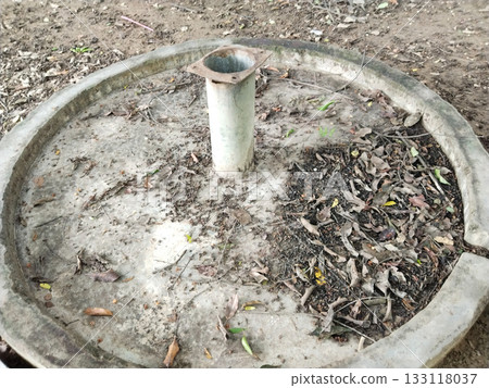 Old concrete water handpump base in park, circular structure with pipe and drainage, 133118037