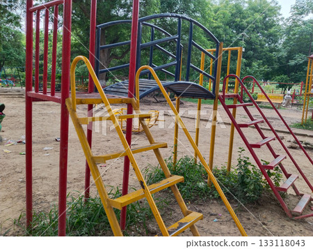 Colorful metal playground structure with ladders, yellow and green slides, climbing arches, and shaded platforms in outdoor park setting for children 133118043